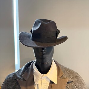 May include: A dark gray fedora hat with a black ribbon band and a small bow on the side. The hat has a wide brim and is displayed on a mannequin wearing a gray blazer and white shirt. The hat is a classic accessory.