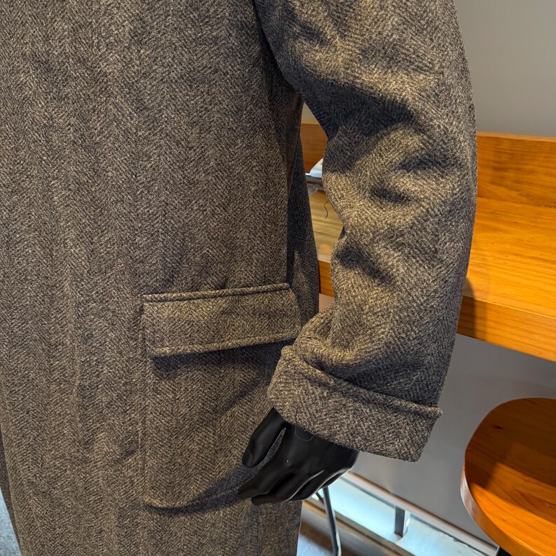May include: A gray herringbone-patterned coat with a pocket on the front. The coat's sleeve is cuffed at the wrist. The person is wearing black gloves. The coat is made of a thick, textured fabric.
