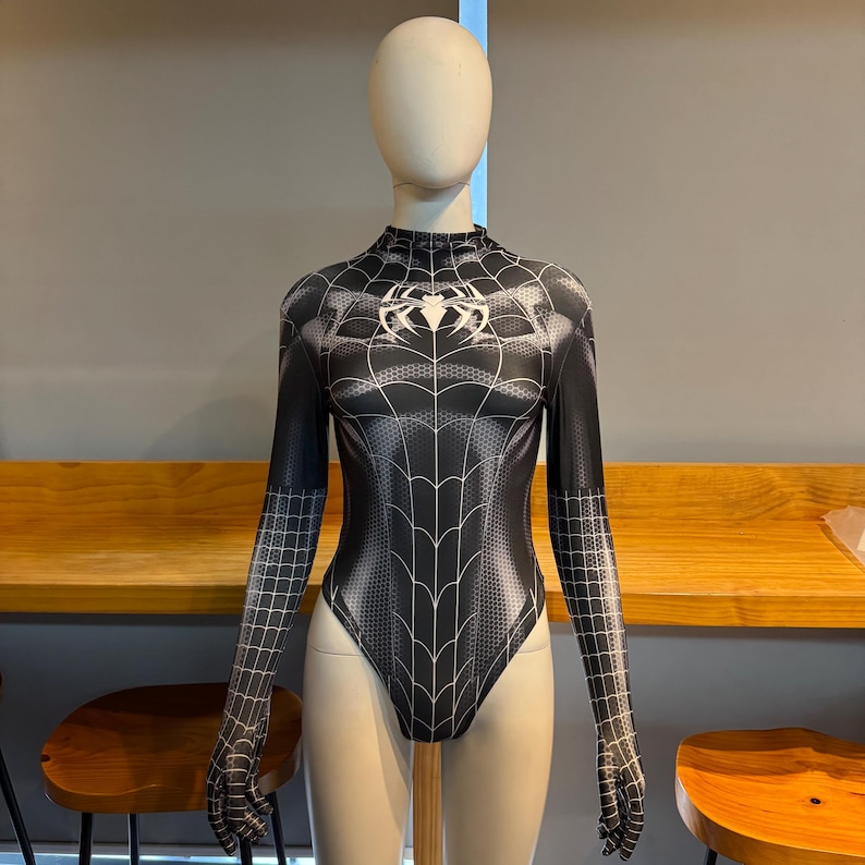 May include: Black bodysuit featuring a spiderweb pattern and a spider emblem on the chest. The long-sleeved garment has a high neckline and a form-fitting shape. The bodysuit is displayed on a mannequin.