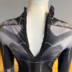 May include: Black and grey jumpsuit with a zip fastening. The fabric has a geometric pattern with hexagonal shapes and web-like designs. The garment is displayed on a mannequin.
