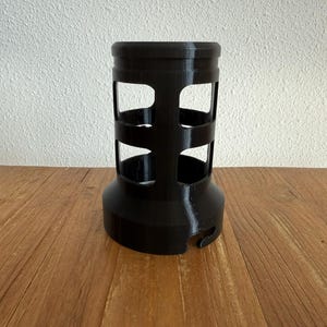 May include: A black, cylindrical object with a series of rectangular cutouts. The object has a wide base and a slightly narrower top, with horizontal bands encircling the cutouts. It is made of a solid, matte material.