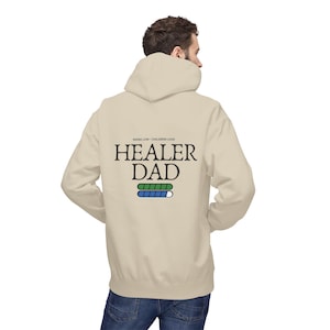May include: A cream-coloured hoodie featuring the text "HEALER DAD" in black, with a green and blue loading bar design. The hoodie also has the text "MANA LOW - CHILDREN LOUD" above the main text. The person is wearing blue jeans.