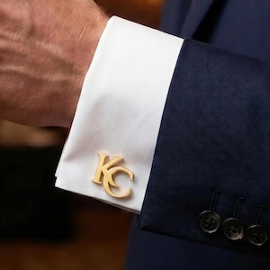 May include: A gold-toned metal cufflink featuring the letters KC in a stylized font, attached to a crisp white French cuff of a dress shirt. The shirt is worn under a dark navy blue suit jacket with four buttons visible on the sleeve cuff.