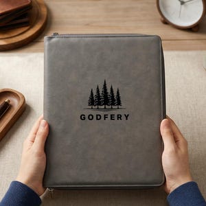May include: A gray zippered portfolio with a black tree graphic and the word "GODFERY" is held by a person. Other items on the table include a pocket watch, a pen, a wooden tray, coasters, a clock, and a small ceramic pot.