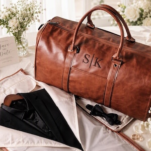 Könnte beinhalten: Eine braune Reisetasche aus Leder mit den eingeprägten Initialen S|K, kombiniert mit einem schwarzen Sakko in einer Kleiderhülle und einer schwarzen Fliege. Die Tasche hat zwei Tragegriffe und eine strukturierte, rechteckige Form.