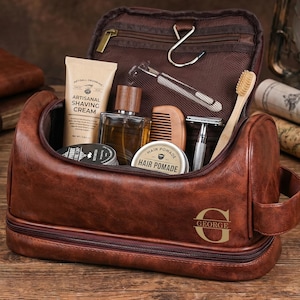 May include: A brown leather toiletry bag with a zipper closure and a handle, featuring the letter G and the name GEORGE embossed on the front. The open bag contains a tube of shaving cream, a glass bottle, a wooden comb, a razor, and hair pomade.