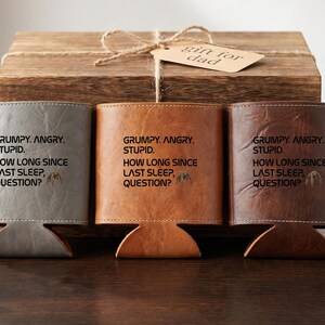 May include: Three leather can coolers in gray, tan, and dark brown, each featuring the text: GRUMPY. ANGRY. STUPID. HOW LONG SINCE LAST SLEEP, QUESTION? with a small bat icon. The items are arranged in front of a wooden box with a gift tag.