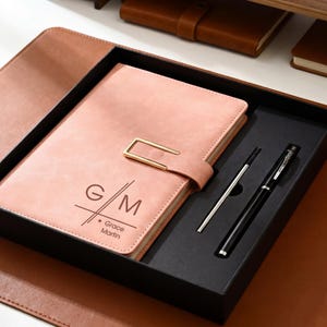 May include: A pink leather journal with a gold clasp and the initials "G/M" and the name "Grace Martin" printed on the cover. The journal is in a black box with a silver pen and a black pen. The set is on a brown leather desk mat.