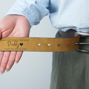 Op de afbeelding: Een bruinleren riem met een donkerbruine leren band en een zilveren gesp. De riem heeft de tekst "HAPPY 1ST FATHER'S DAY AS MY Daddy I LOVE YOU, AMELIA 2026" gegraveerd. De riem wordt door een persoon vastgehouden.