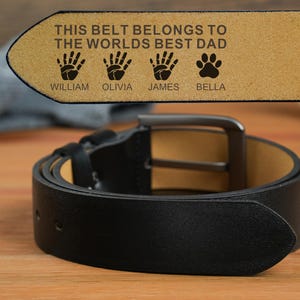 Könnte beinhalten: Ein schwarzer Ledergürtel mit silberner Schnalle, auf einer Holzoberfläche aufgerollt. Das Ende des Gürtels zeigt den Text "THIS BELT BELONGS TO THE WORLDS BEST DAD" mit Handabdrücken, einem Pfotenabdruck und Namen.