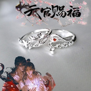 以下が含まれることがあります： 繊細な花のデザインが施された銀の指輪2つ。指輪は絡み合い、小さな赤い石のアクセントが施されています。背景には「天官賜福」の文字が見えます。