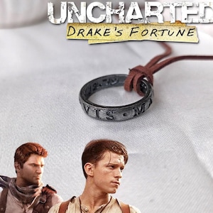 Könnte beinhalten: Eine silberne Ringkette mit den Worten "VIS  MA" eingraviert. Die Kette ist braun und hat eine Lederschnur. Die Kette liegt auf einer weißen Oberfläche mit den Worten "UNCHARTED DRAKE'S FORTUNE" darüber.