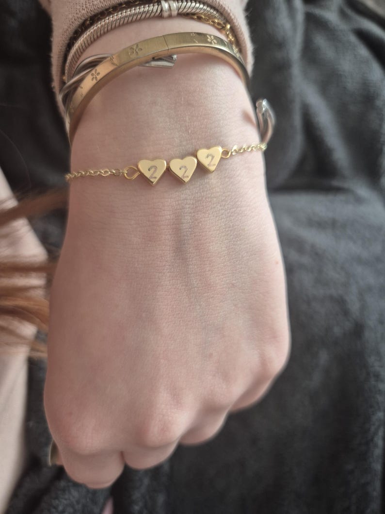 Pu&ograve; includere: Un braccialetto dorato con tre ciondoli a forma di cuore, ognuno con il numero "2". Il braccialetto a catena sottile &egrave; visibile su un polso, insieme ad altri braccialetti dorati. Il design &egrave; semplice ed elegante.