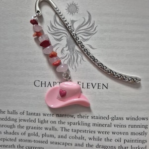 May include: A silver metal bookmark with a decorative design and a pink cowboy hat charm with a red heart. The bookmark also features a strand of pink and red beads. The word "CHAPTER ELEVEN" is visible on the page.