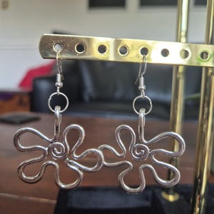 Può includere: Un paio di orecchini a forma di fiore color argento. Ogni orecchino presenta un design ad anello con un centro a spirale e un gancio per orecchie forate. Gli orecchini sono esposti su un espositore per orecchini color oro.