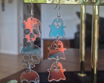 May include: A pair of dangle earrings featuring iridescent acrylic charms. One earring consists of three stacked skull shapes, while the other earring features three stacked ghost shapes. Both earrings are suspended from silver-toned metal hooks.