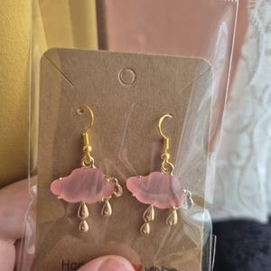 May include: A pair of pink cloud earrings with gold-coloured hooks and teardrop charms. The earrings are presented on a brown card, inside a clear plastic bag. The card has the words "Hand" and "with Love" printed on it.
