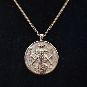 Può includere: Una collana con ciondolo in metallo color oro dal design a moneta circolare. Il ciondolo raffigura una figura a tre teste che regge delle torce, sormontata da un simbolo a mezzaluna. Il pezzo pende da una sottile catena a maglie quadrate.