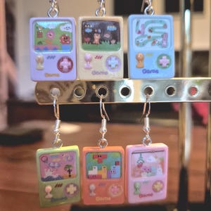 May include: Six dangle earrings shaped like miniature handheld video game consoles in pastel colors including lavender, cream, light blue, lime green, peach, and pink. Each earring features a screen graphic, a joystick, and a directional pad.
