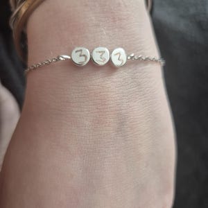 Pu&ograve; includere: Un delicato braccialetto d'argento con tre piccoli ciondoli rotondi. Ogni ciondolo mostra il numero "3" in uno stile semplice e scritto a mano. Il braccialetto &egrave; indossato su un polso, con altri braccialetti visibili sullo sfondo.