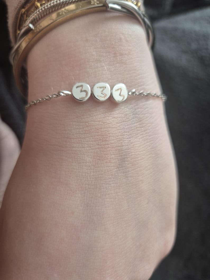 Pu&ograve; includere: Un delicato braccialetto d'argento con tre piccoli ciondoli rotondi. Ogni ciondolo mostra il numero "3" in uno stile semplice e scritto a mano. Il braccialetto &egrave; indossato su un polso, con altri braccialetti visibili sullo sfondo.