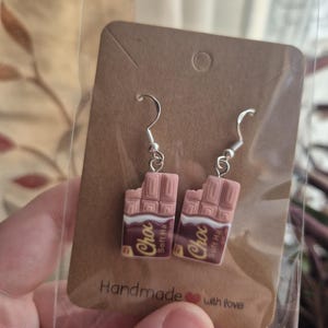 Può includere: Orecchini fatti a mano a forma di barretta di cioccolato. Ogni orecchino presenta un design a barretta di cioccolato rosa con la scritta "Choc" e "Soft Ha". Gli orecchini sono attaccati a ganci per orecchie color argento.