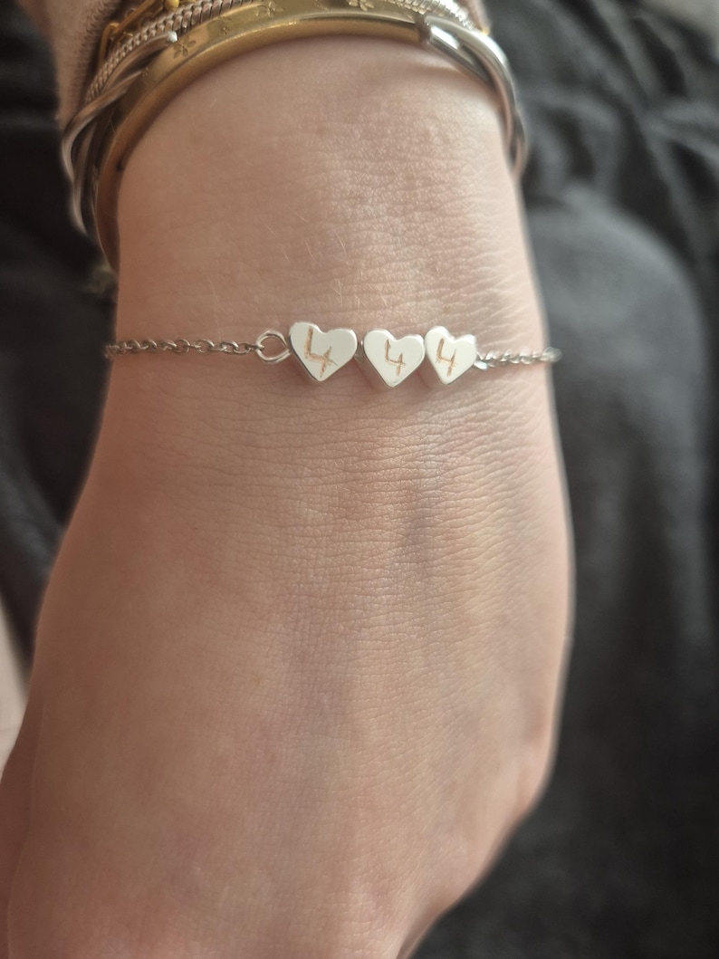 Pu&ograve; includere: Un braccialetto d'argento con tre ciondoli a forma di cuore. Ogni ciondolo ha inciso il numero "4". Il braccialetto &egrave; indossato su un polso, con altri braccialetti visibili sullo sfondo.