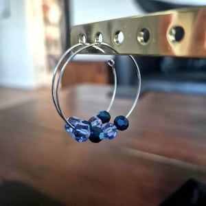 May include: Silver hoop earrings adorned with a cluster of blue beads. The beads range in shade from light to dark blue. The earrings are displayed in front of a gold-coloured metal object.