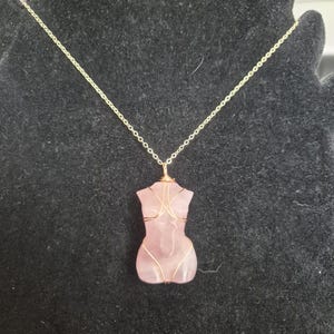 Puede incluir: Un collar con cadena dorada y colgante de piedra rosa. El colgante tiene forma de torso y está envuelto con alambre dorado. El collar se muestra sobre un fondo negro.