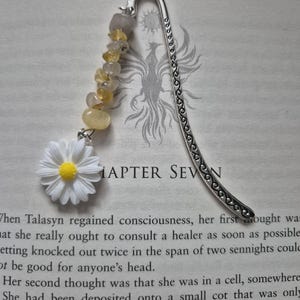 May include: A silver metal bookmark with a daisy charm and yellow gemstone beads. The daisy has white petals and a yellow center. The bookmark is resting on a book with the text "CHAPTER SEVEN".