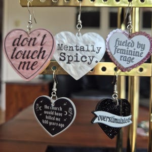 May include: Five heart-shaped dangle earrings with silver-tone hooks. The pink heart says 'don't touch me', the white heart says 'Mentally Spicy.', the glittery heart says 'fueled by feminine rage', the black heart says 'the church would have killed me 400 years ago', and the black glitter heart says 'overstimulated'.