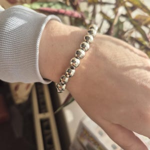 May include: A silver beaded bracelet with faceted, spherical beads. The bracelet is worn on a wrist, with a light grey long-sleeved top visible in the image.