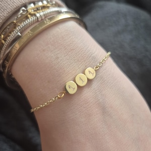 Pu&ograve; includere: Un braccialetto dorato con tre ciondoli circolari, ognuno inciso con il numero "1". Il braccialetto &egrave; delicato, con una catena sottile e un design semplice. Diversi braccialetti d'oro sono impilati sul polso.