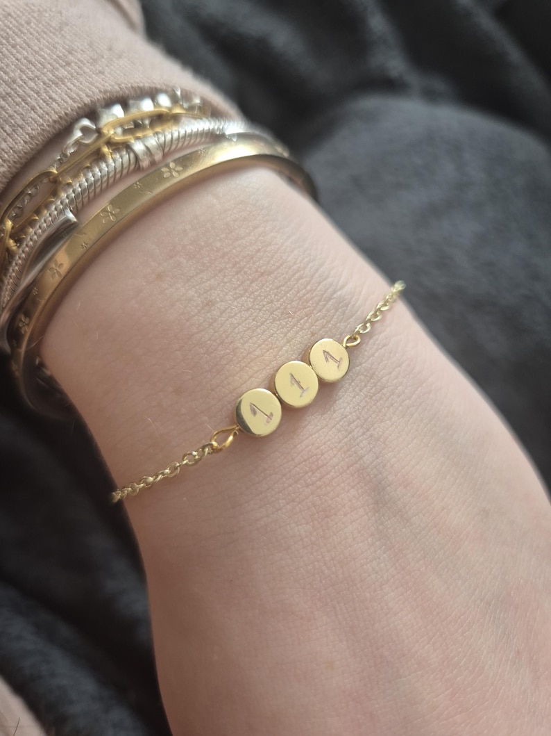 Pu&ograve; includere: Un braccialetto dorato con tre ciondoli circolari, ognuno inciso con il numero "1". Il braccialetto &egrave; delicato, con una catena sottile e un design semplice. Diversi braccialetti d'oro sono impilati sul polso.