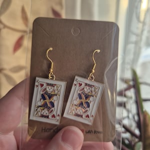 May include: Gold-tone drop earrings with a playing card motif. The cards show a rabbit in regal clothing. The earrings are presented on a brown card with the words "Hand" and "with love".