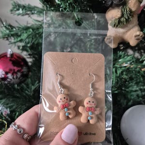 May include: A pair of gingerbread man earrings with silver hooks, displayed on a brown card. The gingerbread men have red bow ties and blue buttons. The card is in a clear plastic bag. The background includes a Christmas tree and a dog figurine.