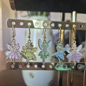 Puede incluir: Cinco pares de pendientes colgantes con temática de hadas. Cada pendiente presenta un diseño de hada colorido con alas, vestidos y ganchos de metal dorado. Las hadas son en tonos rosa, amarillo, verde, azul y morado.