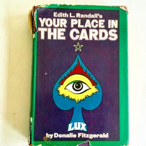May include: A rectangular book titled Edith L. Randall's YOUR PLACE IN THE CARDS by Donalie Fitzgerald. The cover features a purple background with a green border, a central blue spade icon containing an eye, a star, and the word LUX.