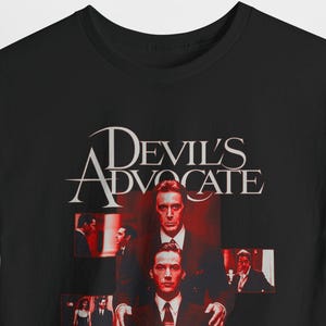 May include: Black t-shirt featuring a movie poster design for "Devil's Advocate." The design includes the movie title in white and red, along with images of actors in suits. The shirt is made of a soft material.
