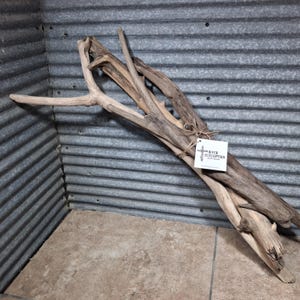 May include: A bundle of weathered driftwood branches with a light brown and grey texture. The branches are tied together with a thin brown twine string. A white square tag is attached to the twine, featuring the text River Redemption Artworks.