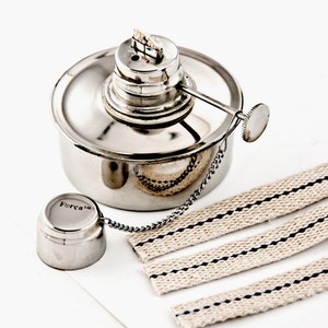 Puede incluir: Una lámpara de alcohol de metal plateado con una mecha y una cadena unida a una pequeña taza de plata que dice "Foreca". También se incluyen tres mechas de algodón blanco con rayas negras.