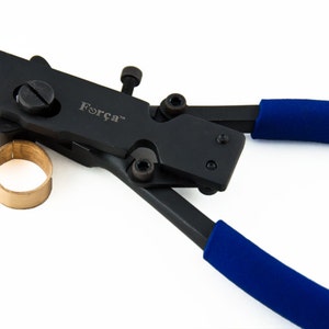 May include: A black metal crimping tool with blue rubberized handles and the brand name Força printed on the side. The tool features multiple adjustment screws and is positioned next to a small, circular, metallic brass-colored ring component.