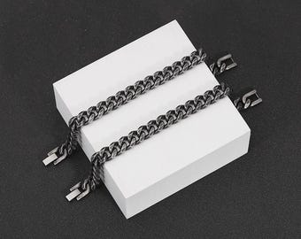 Puede incluir: Dos pulseras de cadena de eslabones plateadas con cierres rectangulares se exhiben sobre un bloque rectangular blanco sobre un fondo oscuro. Las pulseras tienen un diseño de eslabones gruesos e interconectados.