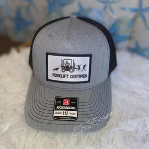 May include: Grey and black trucker cap with a white patch displaying a forklift and the text "Forklift Certified." The cap has a curved peak and an adjustable strap. The Richardson brand label is visible.