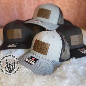 May include: A collection of trucker hats in various shades of gray and black, each featuring a leather patch with the words "DAD" or "DAD in the streets, DADDY in the sheets" in a serif font. The hats have mesh backs and curved brims.