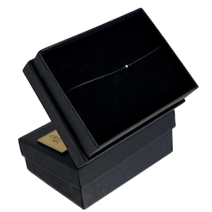 May include: A black jewelry box with a gold-colored accent. Inside, a delicate necklace with a small, gold-colored bead is displayed against a black velvet lining. The box is open, revealing the necklace.