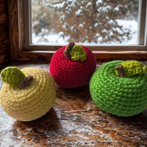 May include: Three crocheted apples in yellow, red, and green sit on a brown surface. Each apple has a brown stem and a green leaf. The background shows a window with a snowy outdoor scene.