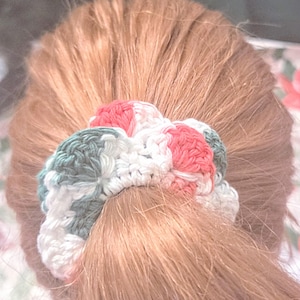 Puede incluir: Una scrunchie de ganchillo hecha a mano en tonos blanco, coral y verde agua. La scrunchie está envuelta alrededor de una coleta de cabello castaño claro. El patrón de ganchillo es visible, mostrando la textura y las variaciones de color.