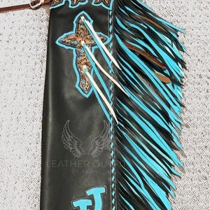 Peut inclure: Chaps en cuir noir avec franges turquoise et noires. Comprend une croix, des coutures décoratives et les initiales "J" et "R" en turquoise. Comprend une sangle de ceinture en cuir.