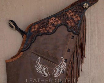 Op de afbeelding: Bruine leren chaps met franjes en bloemmotief. De chaps hebben een zwarte leren bies en een bruine leren riem met een gesp. De chaps zijn versierd met een bruin leren vleugelontwerp en de tekst "Leather Outfit by Abdul".